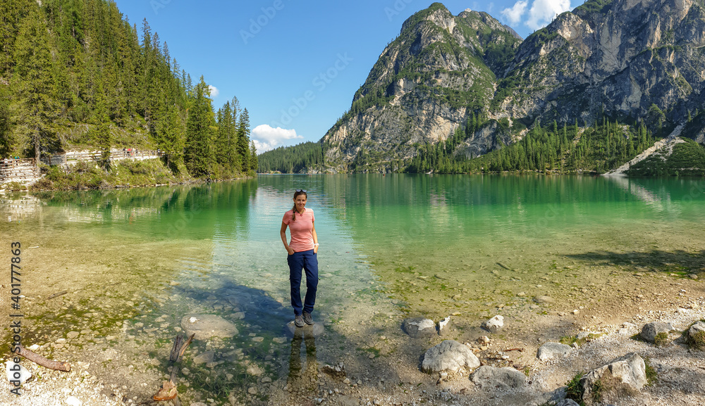 A woman standing on a boulder at the shore of the Pragser Wildsee, a lake in South Tyrolean Dolomites. High mountain chains around the lake. The sky and mountains are reflecting in the lake. Calmness