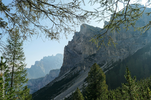 A view on a steep mountain wall in Italian Dolomites. There is another mountain chain behind it. The sunbeams reaching back sides of the mountain. A few tree branches disturbing the clear view
