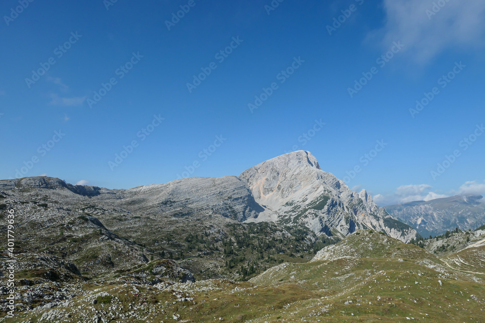 A panoramic view on a vast valley in Italian Dolomites. There are high mountain chains around. The bottom of the valley is lush green. A few trees on the slopes. Few soft clouds on the sky. Freedom