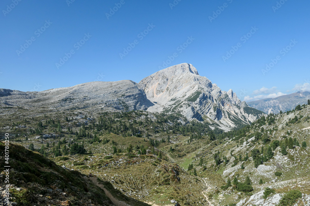 A panoramic view on a vast valley in Italian Dolomites. There are high mountain chains around. The bottom of the valley is lush green. A few trees on the slopes. Few soft clouds on the sky. Freedom