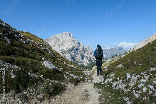 A man with a hiking backpack hiking on a narrow pathway in high Italian Dolomites. Steep  sharp mountain chain in the back. She is crossing a lush green meadow. Discovering and exploring the nature