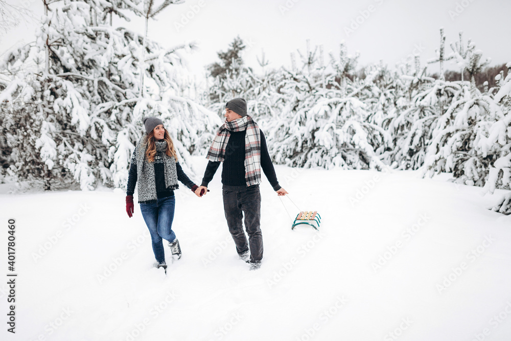 A loving couple walks with a sled, holding hands in winter in a snowy forest with a sled, looking at each other