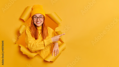 Look at this amazing offer. Cheerful young female model has red hair toothy smile points right on blank space advertises some item dressed in yellow sweater jumper eyeglasses poses in ripped hole © wayhome.studio 