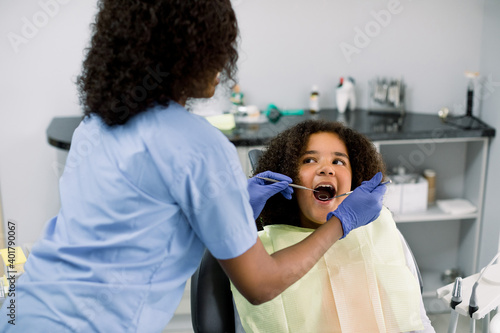 Close up of pretty mixed raced teen girl in dentist chair  with mouth open  having dental examination. Back view of female African dentist in gloves  holding dental tools