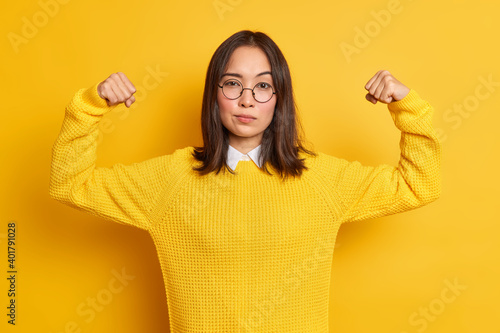 Confident young Asian woman shows arm muscles feels like hero demonstrates her power and strength looks seriously at camera wears round optical glasses sweater isolated over yellow background