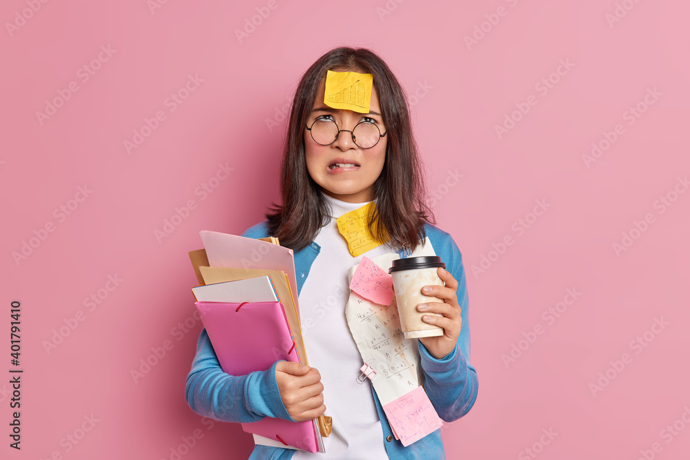 Displeased university student bites lips and looks above with unhappy tired expression drinks takeaway coffee works with papers wears round spectacles isolated over pink background has deadline