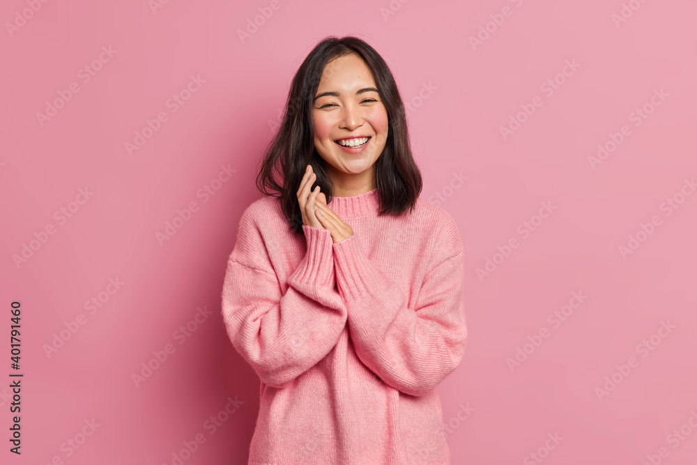 Fototapeta premium Portrait of sincere brunette Asian female model rubs palms and smiles broadly expresses happiness feels joyful has white even teeth wears oversized casual sweater isolated over pink background
