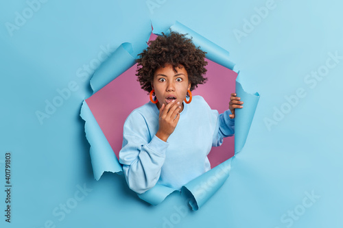Billede på lærred Shocked dark skinned Afro American woman stares at camera holds breath from wonder keeps mouth opened wears casual jumper breaks through paper background sees something unbelievable
