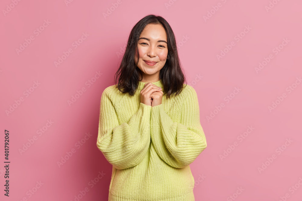 Pretty young Asian woman keeps hands together smiles pleasantly glad to receive compliment from boyfriend dressd in knitted yellow sweater isolated on pink background. Pleasant human emotions concept Pretty young Asian woman keeps hands together smiles pleasantly glad to receive compliment from boyfriend dressd in knitted yellow sweater isolated on pink background. Pleasant human emotions concept