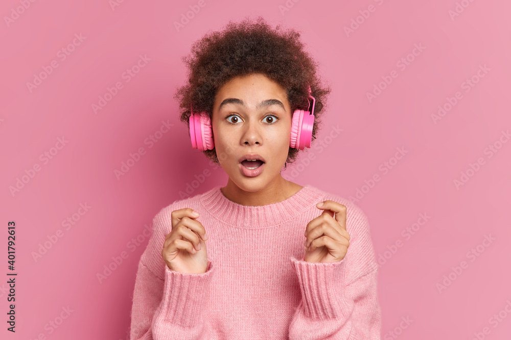 Shocked curly haired pretty millennial girl stares at camera gasps from amazement keeps mouth opened reacts on surprising news wears stereo headphones casual jumper isolated over pink background Shocked curly haired pretty millennial girl stares at camera gasps from amazement keeps mouth opened reacts on surprising news wears stereo headphones casual jumper isolated over pink background