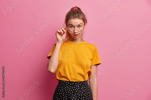 Half length shot of astonished female student cannot believe in her results on exams looks wondered through spectacles wears yellow t shirt and skirt expresses shock and amazement. Reaction concept