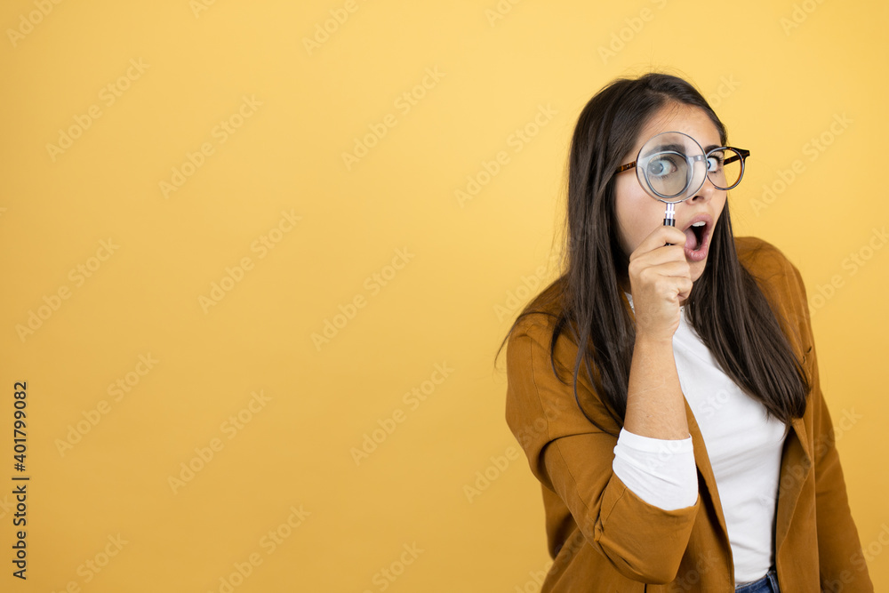 Young beautiful woman wearing a blazer over isolated yellow background surprised looking through a magnifying glass