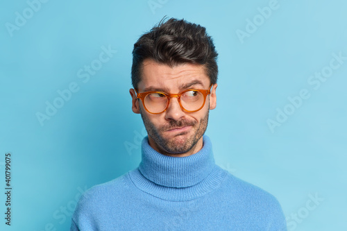 Close up shot of serious thoughtful brunet man has thick bristle purses lips wears transparent glasses and poloneck poses against blue background concentrated aside feels hesitant about something