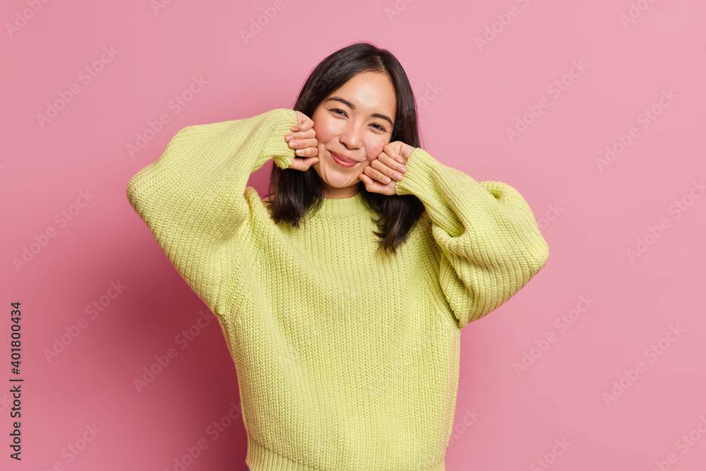 Cute dark haired female model looks pleasantly at camera touches cheeks gently smiles tenderly wears yellow knitted jumper feels glad has natural beauty isolated over pink background. Tenderness Cute dark haired female model looks pleasantly at camera touches cheeks gently smiles tenderly wears yellow knitted jumper feels glad has natural beauty isolated over pink background. Tenderness
