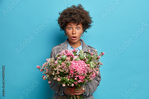Beautiful curly haired woman keeps mouth opened holds bunch of nice flowers reacts on something amazing dressed in jacket poses against blue background. Spring time concept. Female with bouquet