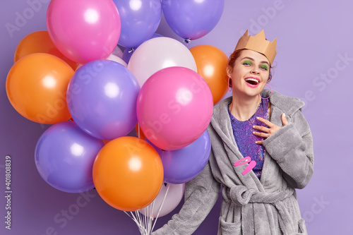 Positive birthday girl feels pleased to receive congratulations from friends dressed in dressing gown handmade corona holds big bunch of multicolored balloons isolated over purple background