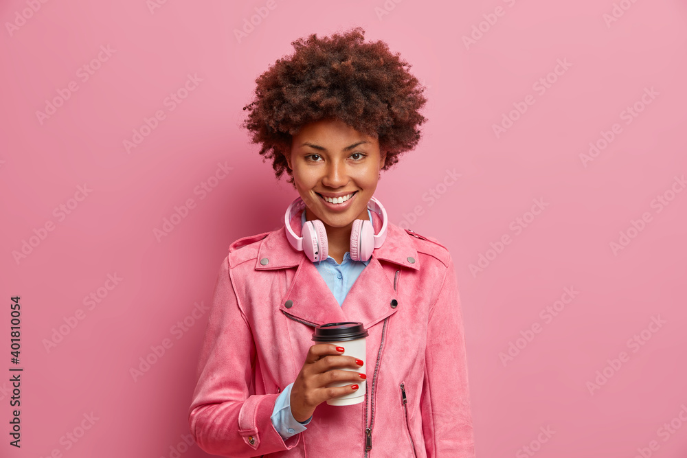 Pleased pretty young African American woman holds disposable cup of aromatic coffee wears headphones around neck dressed in stylish jacket expresses positive emotions isolated over pink background Pleased pretty young African American woman holds disposable cup of aromatic coffee wears headphones around neck dressed in stylish jacket expresses positive emotions isolated over pink background