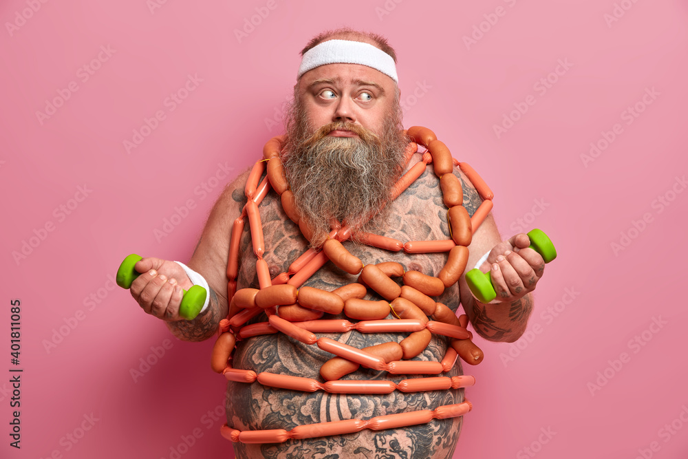 Fat bearded man frustrated because of diet failure poses with two dumbbells sausages around body has problem of overweight tries to go in for sport and loose weight isolated over pink background