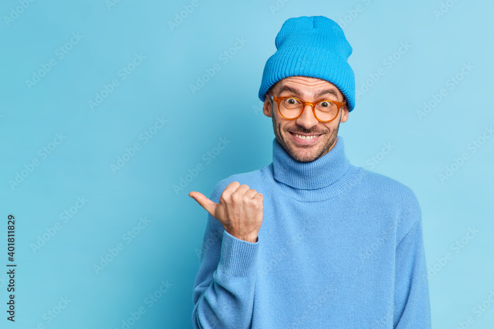 Horizontal shot of cheerful guy with bristle points thumb on left side wears blue hat and turtleneck gives direction to promotion isolated over blue background. People advertisement concept. Horizontal shot of cheerful guy with bristle points thumb on left side wears blue hat and turtleneck gives direction to promotion isolated over blue background. People advertisement concept.