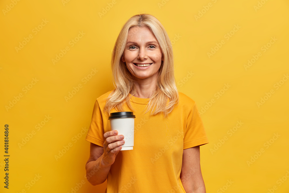 Horizontal shot of blonde European woman with pleasant smile minimal makeup holds disposable cup of coffee dressed in casual t shirt isolated over yellow studio wall. People age lifestyle concept Horizontal shot of blonde European woman with pleasant smile minimal makeup holds disposable cup of coffee dressed in casual t shirt isolated over yellow studio wall. People age lifestyle concept