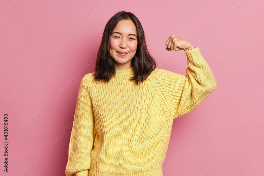 Horizontal shot of pretty Asian woman raises arms shows muscles being strong person has power dressed in casual yellow jumper poses against pink background. Look at my biceps after training. Horizontal shot of pretty Asian woman raises arms shows muscles being strong person has power dressed in casual yellow jumper poses against pink background. Look at my biceps after training.