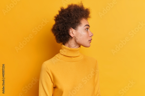 Studio shot of curly woman stands sideways against yellow background turns head aside has serious expression dark curly hair dressed in turtleneck wears round earrings notices something on right © wayhome.studio 