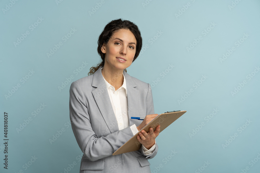 Friendly insurance agent woman wear grey blazer and white blouse holding clipboard, pen looking at camera. Smiling female office worker isolated on studio blue background. Friendly insurance agent woman wear grey blazer and white blouse holding clipboard, pen looking at camera. Smiling female office worker isolated on studio blue background.