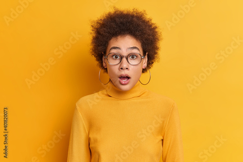 Portrait of shocked curly woman gazes at camera with opened mouth reacts on amazing news wears transparent glasses earrings turtleneck. Speechless impressed Afro American teenager keeps jaw dropped © wayhome.studio 