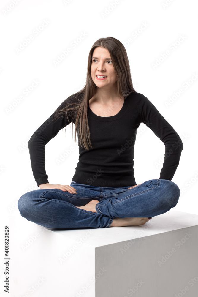 Angry and irritated young woman sitting in the lotus pose on the white cube. Isolated image on the white studio background. Angry and irritated young woman sitting in the lotus pose on the white cube. Isolated image on the white studio background.