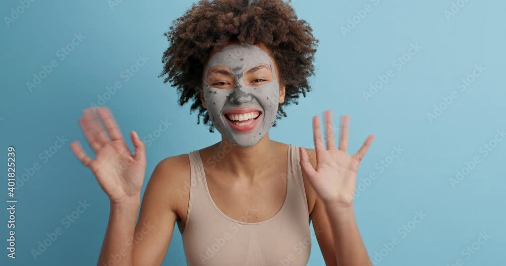 Optimistic dark skinned curly woman appears suddenly smiles gladfully and waves palms greets best friend applies clay mask for skin cleansing and removing wrinkles isolated over blue background Optimistic dark skinned curly woman appears suddenly smiles gladfully and waves palms greets best friend applies clay mask for skin cleansing and removing wrinkles isolated over blue background