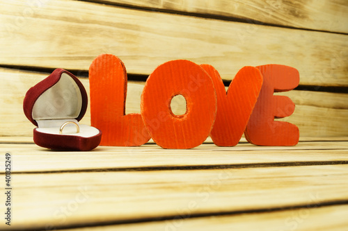 A wedding ring in a box in the shape of a heart, and volumetric letters on a wooden background with copy space.