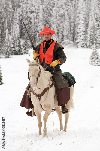 adult cowboy deer hunter holding rifle on white horse in snowy wilderness