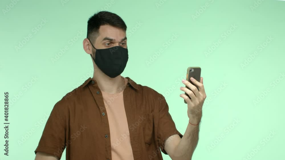 Quarantine, virus and health concept. Cheerful man with dark hair and thick eyebrows, wearing black face mask, video call on smartphone, waving hand at phone camera to say hi Quarantine, virus and health concept. Cheerful man with dark hair and thick eyebrows, wearing black face mask, video call on smartphone, waving hand at phone camera to say hi
