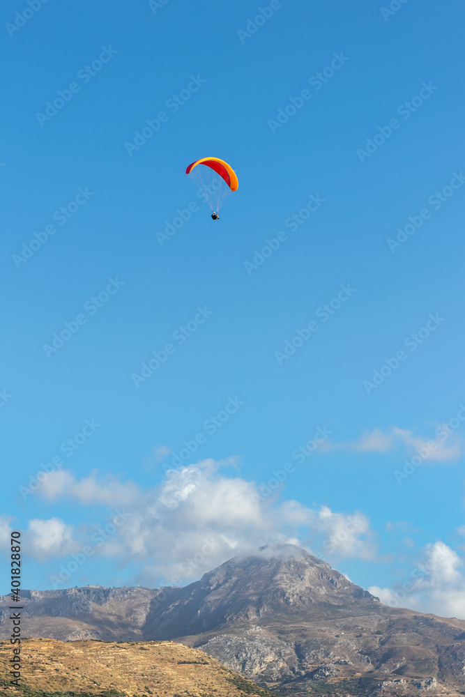 Paragliding in an orange paraglider against a clear blue sky in a bright summer’s day in Crete, Greece. Hills and mountain with small clouds in the background. Paragliding in an orange paraglider against a clear blue sky in a bright summer’s day in Crete, Greece. Hills and mountain with small clouds in the background.