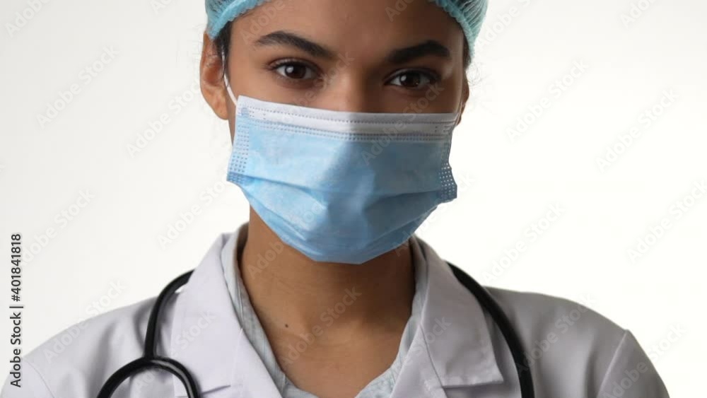 Receive care. Close up of attractive young female doctor wearing medical mask looking at camera, posing isolated on white background. Healthcare and medicine concept. Camera is moving up, slow motion Receive care. Close up of attractive young female doctor wearing medical mask looking at camera, posing isolated on white background. Healthcare and medicine concept. Camera is moving up, slow motion