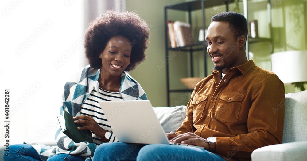 Family couple together resting at home. Pretty African American woman sitting on sofa and reading enjoying favorite book while her husband typing and browsing on laptop. Marriage concept - obrazy, fototapety, plakaty 