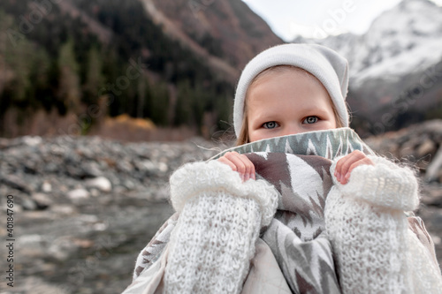 Portrait of preteen blond girl dressed in knitted warm hat, sweater and wrapping plaid blanket standing by the river . Happy hiker wanderlust the view of snow, mountains