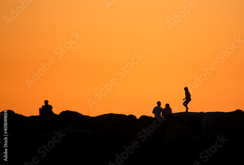 Silhouette against an orange sky of a family of three and a separate individual evoking emotions of family versus isolation during sunset at the Corona del Mar beach in Orange county, California.