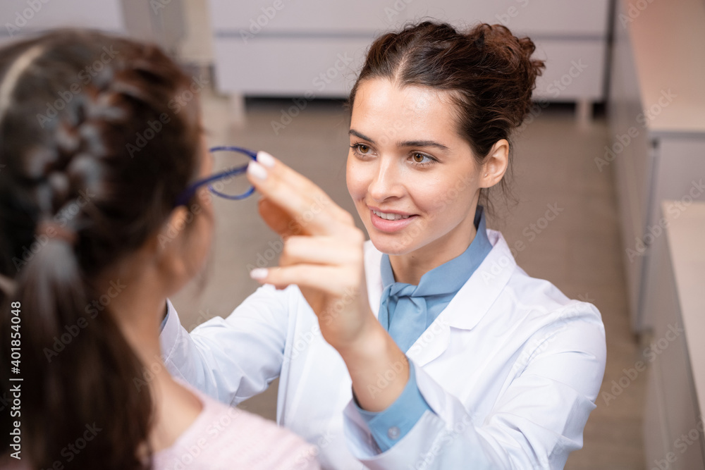Happy ophthalmologist putting pair of new eyeglasses on face of cute little girl Happy ophthalmologist putting pair of new eyeglasses on face of cute little girl