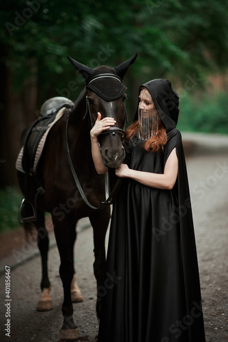 Young woman in hat and black cloak with a horse in the forest