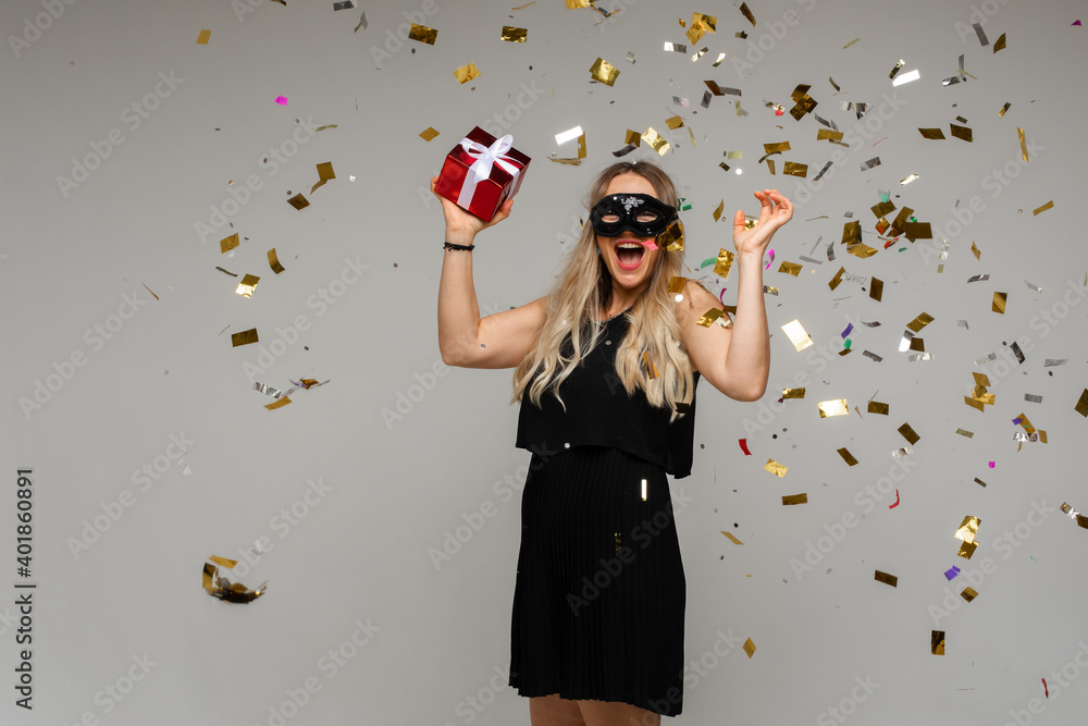 Fototapeta premium Cheerful blonde woman in black cocktail dress and masquerade mask holding arms up with a red Christmas gift having fun under flying confetti. New Year’s night party.