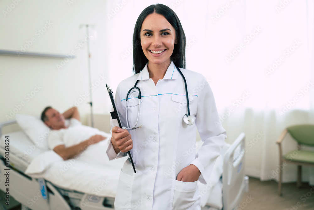 Close up photo of a smiling confident cute woman doctor in a coat with a stethoscope is looking on the camera with the patient in bed in the background Close up photo of a smiling confident cute woman doctor in a coat with a stethoscope is looking on the camera with the patient in bed in the background
