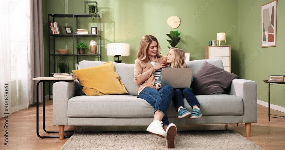 Young Caucasian mother and daughter using laptop for education sitting on sofa in home during quarantine. Beautiful woman and child looking at computer screen with happy smiles browsing online Young Caucasian mother and daughter using laptop for education sitting on sofa in home during quarantine. Beautiful woman and child looking at computer screen with happy smiles browsing online