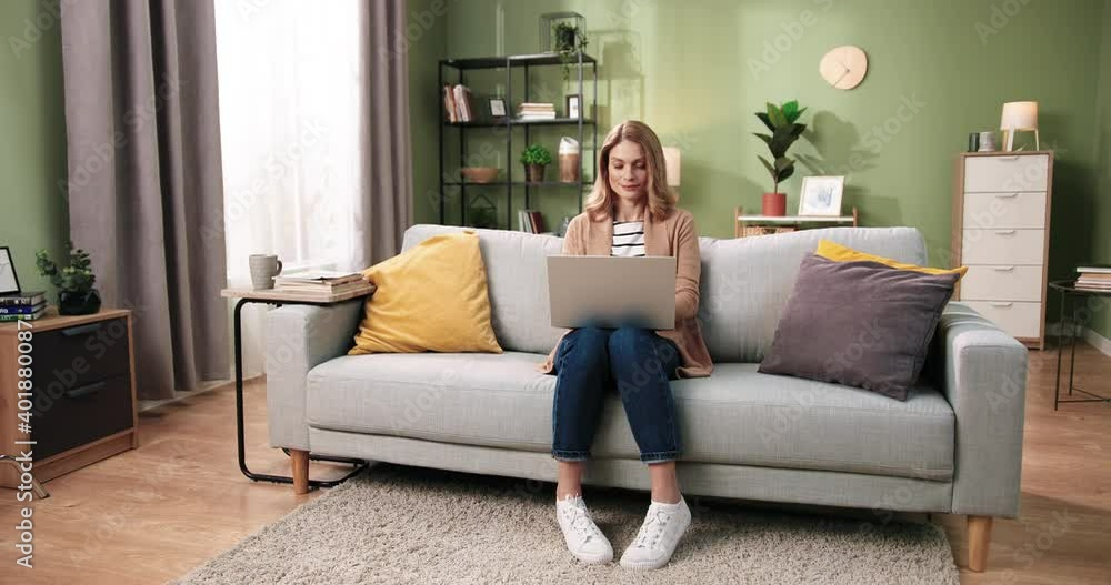 Caucasian beautiful young woman sitting on sofa in cozy living room in apartment texting and working on laptop. Distance work from home. Leisure concept. Browsing online on computer Caucasian beautiful young woman sitting on sofa in cozy living room in apartment texting and working on laptop. Distance work from home. Leisure concept. Browsing online on computer