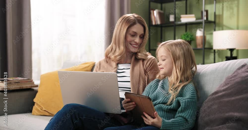 Portrait of joyful smiling Caucasian young mother working and texting on laptop sitting with little cute daughter browsing online on tablet device, speaking and spending time together at home on sofa Portrait of joyful smiling Caucasian young mother working and texting on laptop sitting with little cute daughter browsing online on tablet device, speaking and spending time together at home on sofa