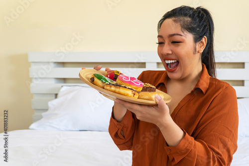 Unhealthy eating concept. Overweight fat woman holding plate with doughnut while sitting on the bed at home. Plus-size fatty girl from sweet donut and bad junk food.