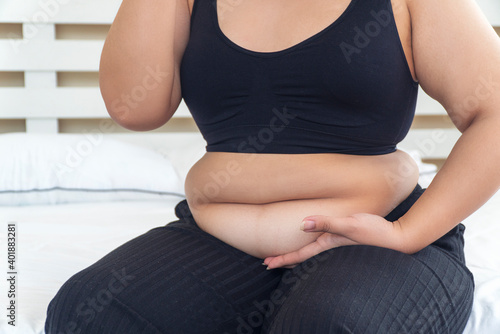 Overweight young woman pinches fat on her belly large size while sitting on white bed at home. Upset female suffering from extra weight. Obesity unhealthily concept. photo