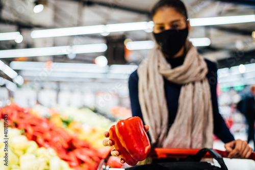 Young woman wearing protective face mask shopping in a supermarket,buying organic produce and ingredients.Eating healthy food during coronavirus COVID - 19 pandemic.Healthy bell pepper.Grocery shop