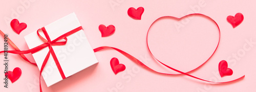 Heart shape made of red ribbon and white gift box with red bow on pink background. Card for Valentines, Mother, Women's day and 8 March. Copy space, mock up, banner photo