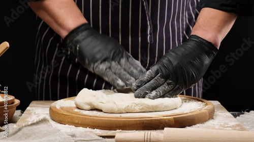 chef in black latex gloves kneading white wheat flour dough on a round wooden board, preparing pizza dough